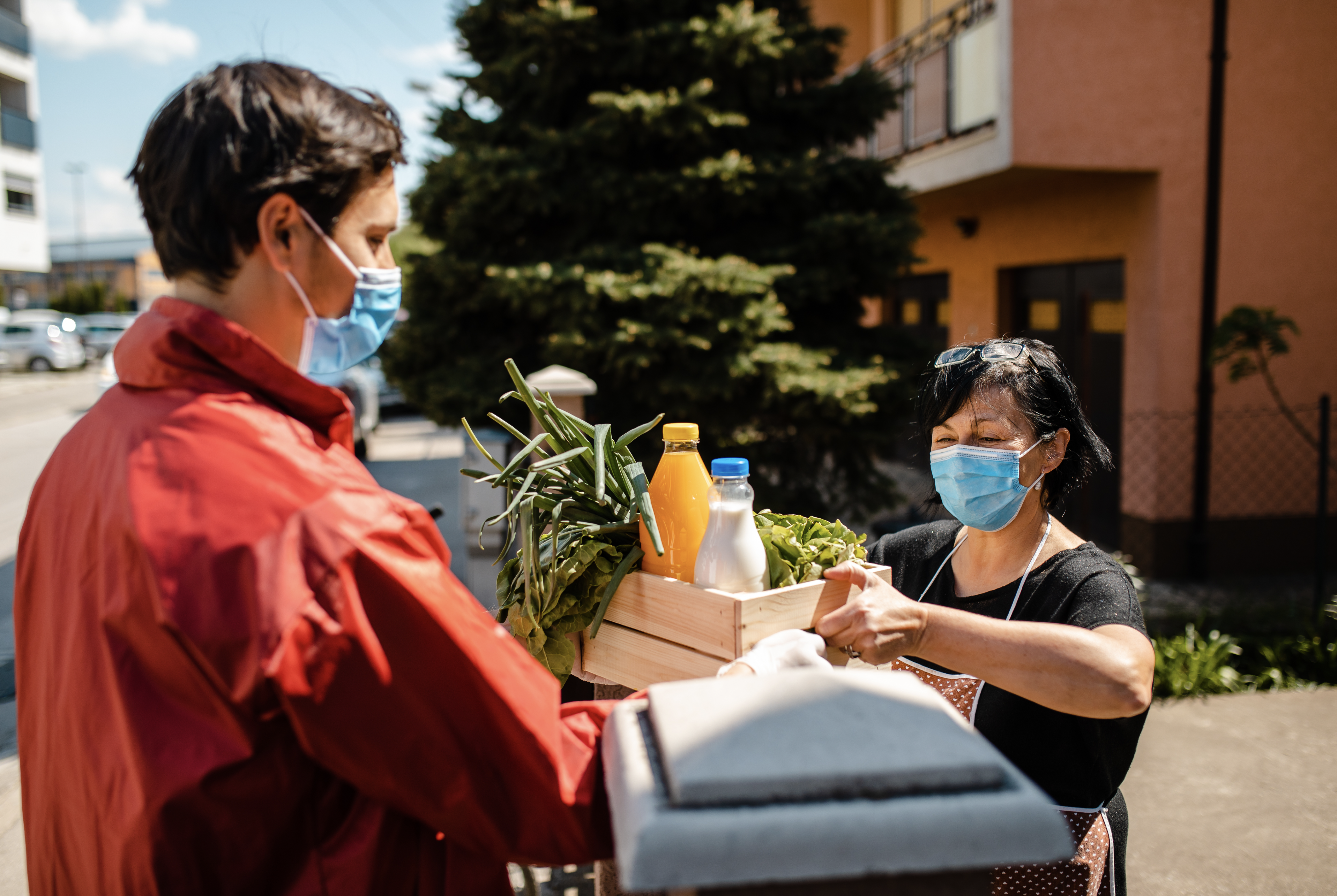 Young male delivery person delivering a crate with fresh vegetables to a senior woman, while they are wearing protective gloves and face masks during the coronavirus pandemic